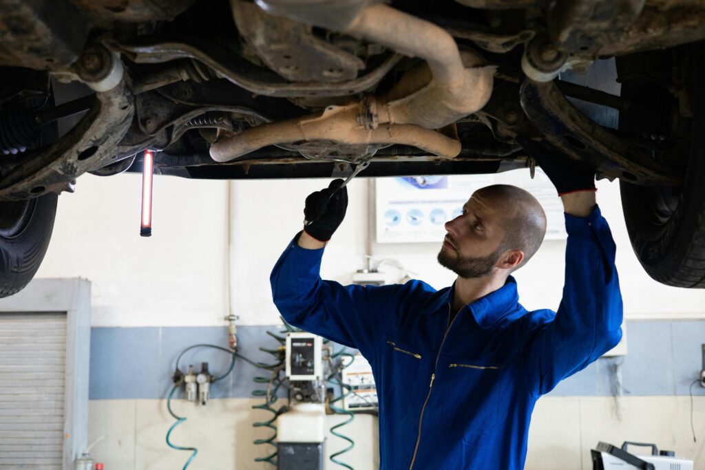 A mechanic in blue overalls inspects the exhaust system of a car in a workshop.